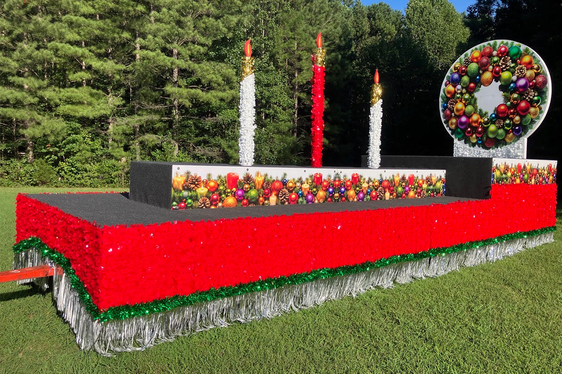 A Christmas parade float featuring three tall decorative candles and a large circular Christmas wreath decorated with colorful ornaments.